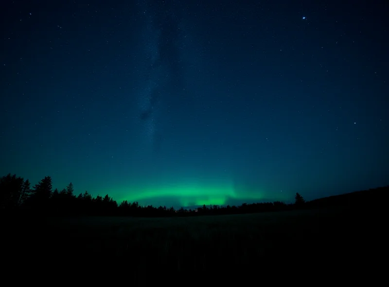 A wide-angle landscape photo of a starry night sky above a field, with a faint green glow from the aurora borealis in the distance.